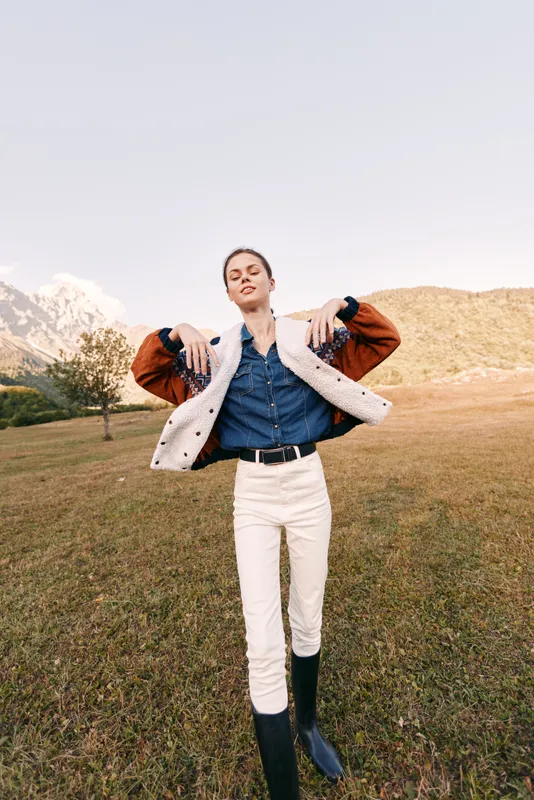 Woman fashion model outdoors in mountains wearing shearling jacket and denim shirt with white pants and black boots, posing confidently on grassy field for lifestyle editorial photography.