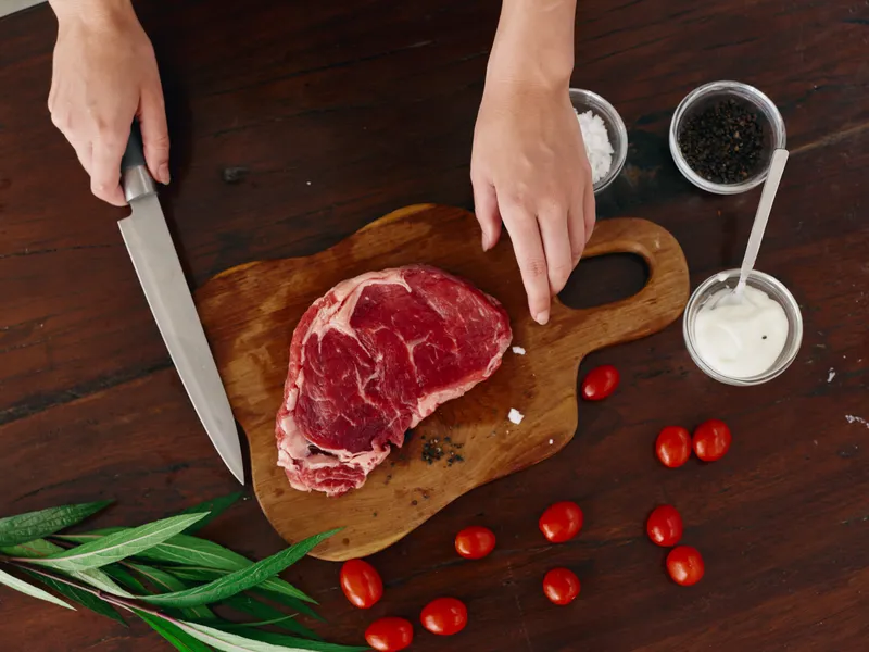 Женщина с ножом в руке нарезает свежее мясо стейка для жарки на кухне