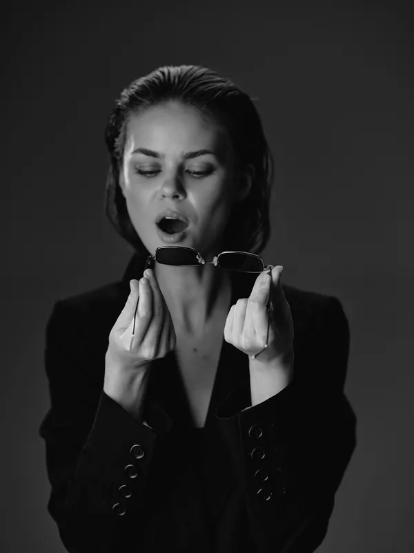 Surprised woman in black blazer holding sunglasses with open mouth in black and white portrait, expressive emotion and formal style.