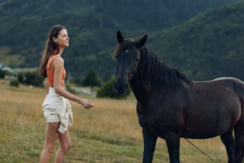A young woman in a sunlit field shares a gentle moment with a horse, smiling softly as they connect in a peaceful rural setting.
