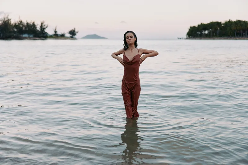 A woman stands in shallow sea water, wearing a red jumpsuit, with hands on hips and a composed expression, as soft light glows over a calm shoreline.