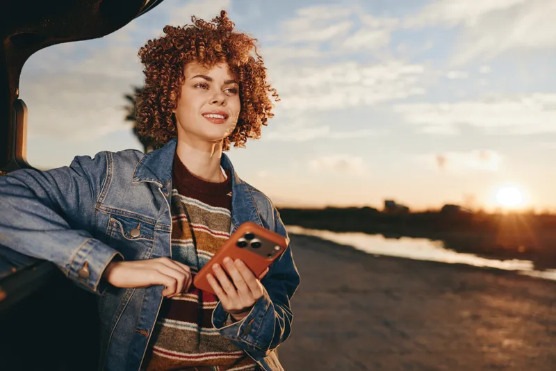 Woman with curly hair, denim jacket, holding smartphone for selfie at beach sunset, cinematic golden light, relaxed lifestyle mood, outdoor casual portrait, near ocean waves, travel today
