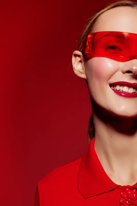Close-up of smiling woman wearing red transparent glasses and red shirt against red background. Concept of fashion, style, beauty, and modern accessories with vibrant color theme.