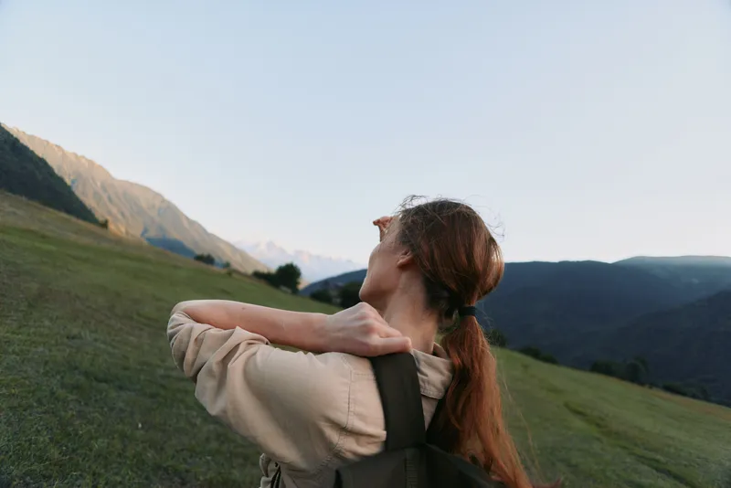 Young woman with backpack stands on a grassy hillside as distant mountains glow softly, inviting a moment of calm and freedom.