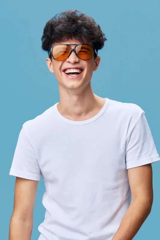 Smiling man with sunglasses wearing a white t-shirt on blue background, expressing happiness and confidence with curly hair, casual style, summer vibes.