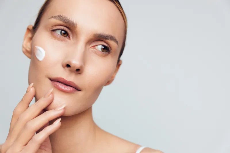 A young woman with clear skin applies white facial cream on her cheek with fingers. The close-up beauty portrait shows skincare and natural complexion on a light background.