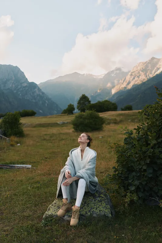 Woman sits on a rock in a meadow with mountains in the background, wearing a cozy cardigan and boots, enjoying peaceful outdoor scenery and fresh air.