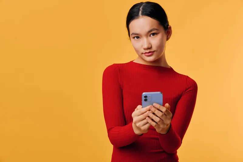 Woman in red sweater holding smartphone with both hands looking at camera with slight smile against yellow background. Female person using mobile device indoors for communication or social media.