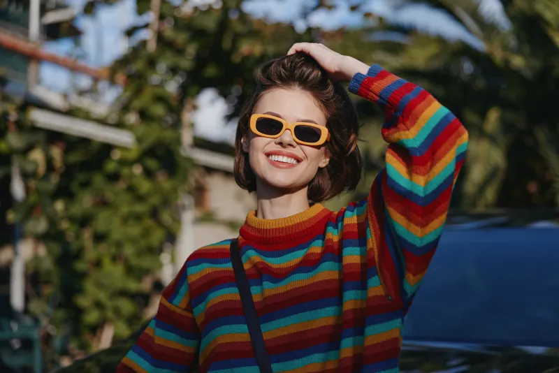 Woman smiling in a colorful striped sweater wearing yellow sunglasses, short hair and casual outfit outdoors among palm trees, relaxed travel vibe and sunny lifestyle.