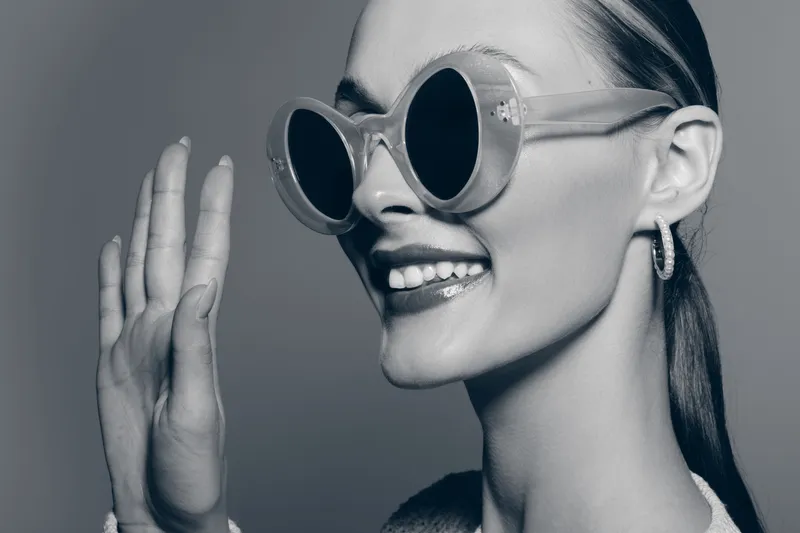Smiling Caucasian woman with pale skin wears large round sunglasses and hoop earrings, showing hand gesture. Studio portrait with retro fashion concept in black and white.