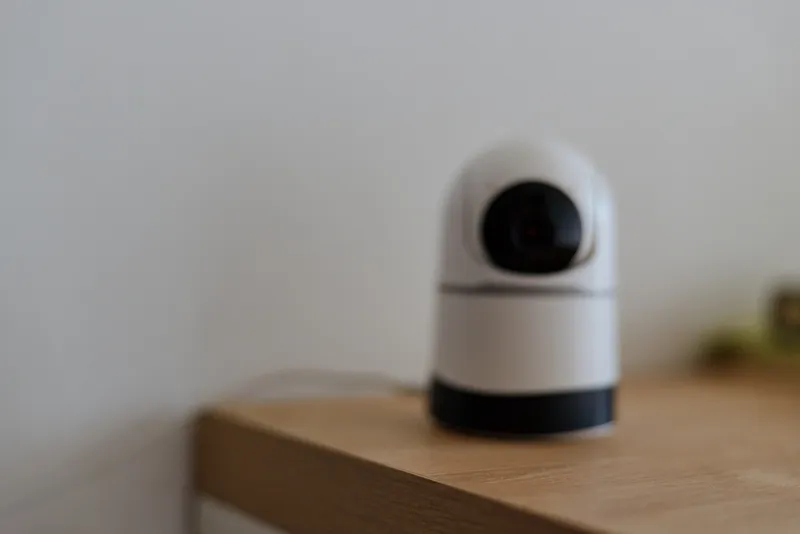A compact home security camera sits on a wooden desk, watching a quiet room with a calm, vigilant presence and a hint of modern technology in a domestic setting.