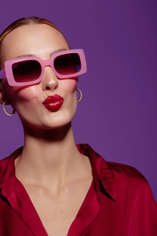 Stylish woman with pink sunglasses and red lipstick makes a kiss face on purple background. Fashion portrait with bold makeup and trendy accessories in studio lighting.