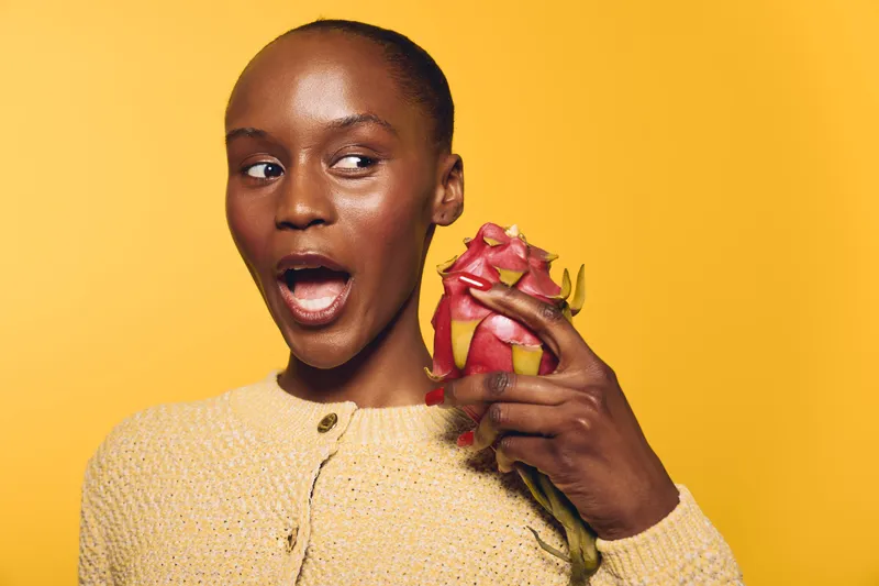 Surprised African woman with dark skin holds dragon fruit near face on yellow background. Concept of exotic fruit, healthy eating, and natural beauty with vibrant studio lighting.