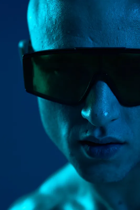 Close-up portrait of a man in sunglasses, illuminated by vibrant blue lighting, showcasing a dramatic and intense expression that captures a futuristic vibe.