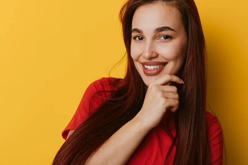 Young woman with long hair smiling and posing on yellow background wearing red shirt, beauty portrait with confident expression and natural makeup for lifestyle and happiness.