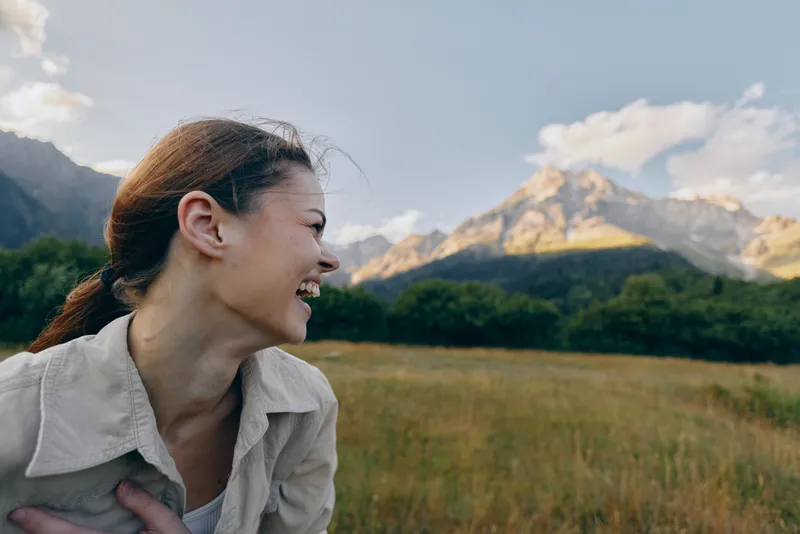 Happy woman with ponytail laughs outdoors in nature with mountains and blue sky in background, enjoying lifestyle and fresh air in sunny day.