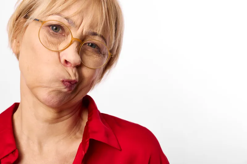 Woman portrait glasses senior blonde expression red studio closeup, close framed face with short hair, golden eyewear, red shirt, neutral studio light, calm playful pout, soft shadows