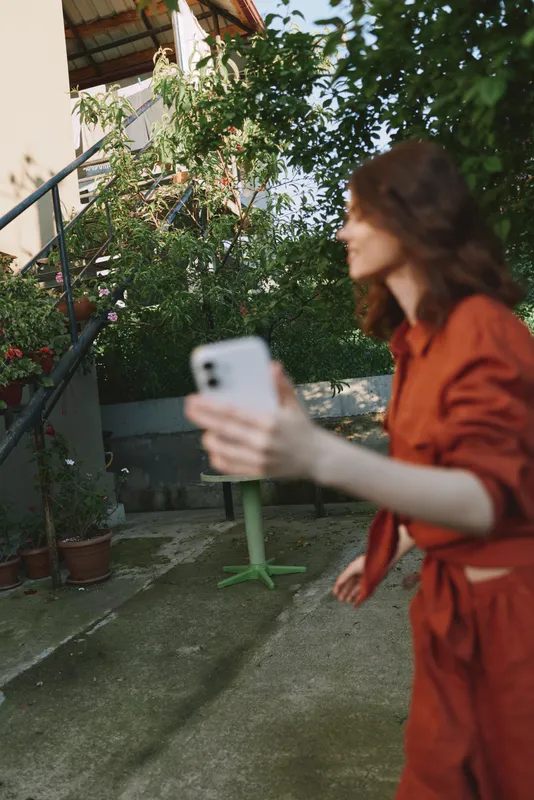 Woman lifestyle on a sunlit balcony, wearing a rust red outfit, holds smartphone for a casual selfie, candid moment, soft focus background, relaxed pose