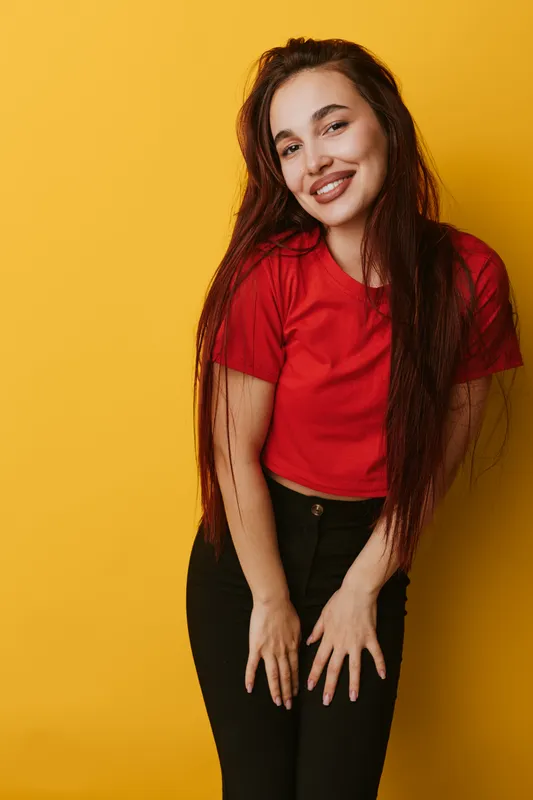 Young woman smiling and posing in casual red shirt and black pants against yellow background. Portrait of happy female with long hair and relaxed posture in studio lighting.