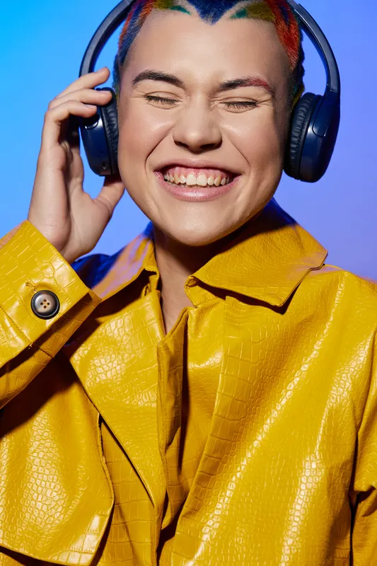 Young woman with colorful hair smiling joyfully while listening to music on headphones, wearing a bright yellow jacket against blue background.