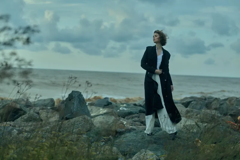 Woman in a long dark coat stands on a rocky shoreline, windswept and serene, with the ocean and cloudy sky behind, conveying fashion, nature, and a calm coastal mood for stock photography
