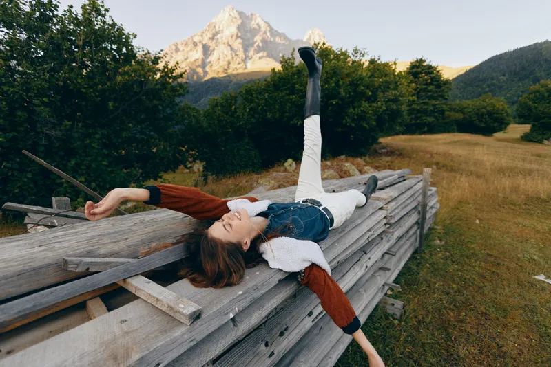 Woman relax countryside on wooden fence with legs raised, casual outfit and knit sweater in meadow, distant mountains at golden hour, outdoor leisure and carefree rural resting portrait.