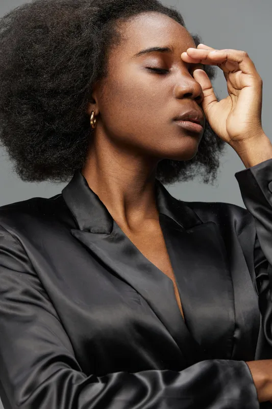 Elegant African woman in a black satin suit expressing deep thought against a neutral gray background, showcasing emotion and contemporary fashion