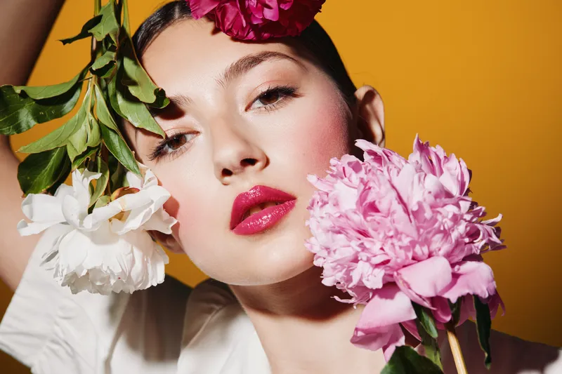 Fresh woman with pink lips posing creatively with white and pink flowers in studio against warm yellow background. Concept for beauty, fashion, creative, skincare.