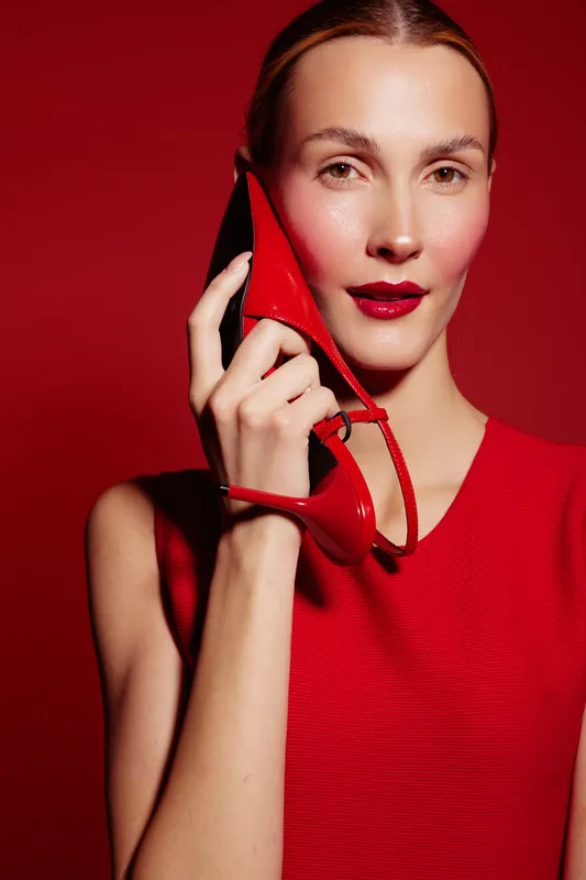 Young woman in red dress holds red high heel shoe near face against red background. Fashion concept with bold makeup and stylish accessories in vibrant studio lighting.