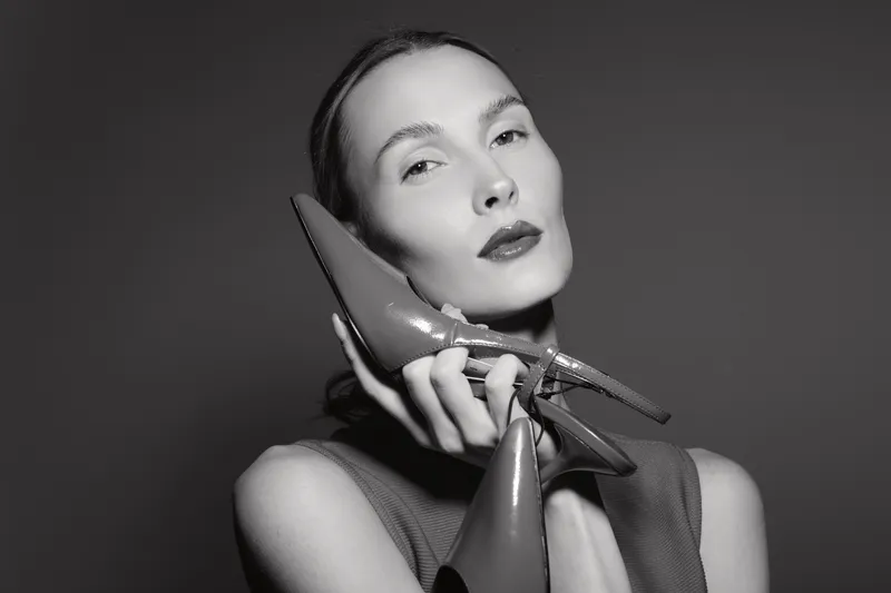 White caucasian woman with pale skin and dark hair holds shiny high heel shoes near face in stylish black and white portrait. Concept of fashion, elegance, and beauty.