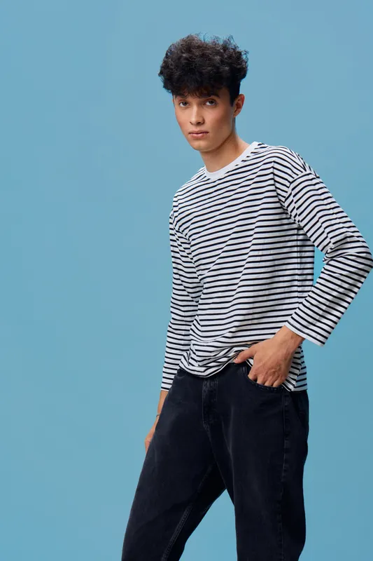 Young man with curly hair wearing striped long sleeve shirt and black pants standing against blue background in casual confident pose.