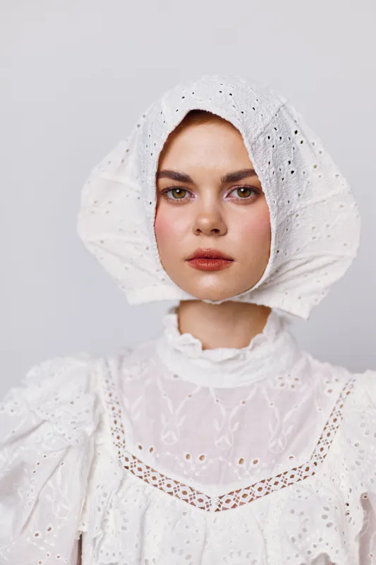 Young woman in a white vintage dress and bonnet, showcasing a serene expression against a soft gray background, highlighting beauty and simplicity