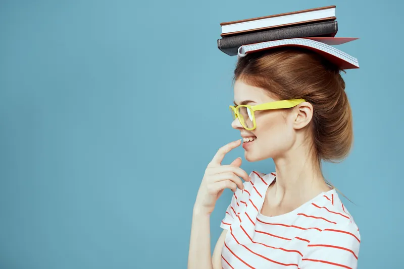 Студентка держит книги на голове и в жёлтых очках на синем фоне, обрезанный вид