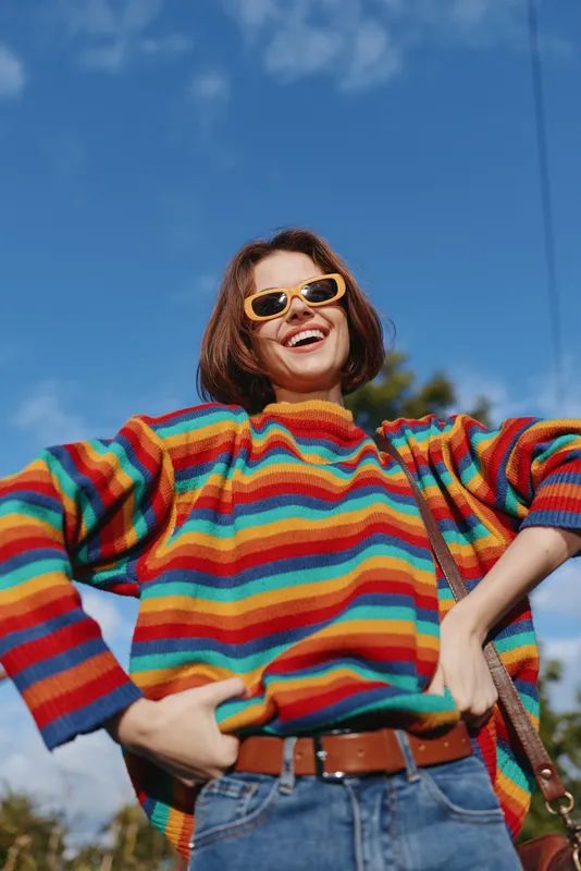 Woman, rainbow sweater, sunglasses, smiling, casual outfit, outdoors: young woman in her 20s with short brown hair wearing rainbow striped sweater and yellow sunglasses, smiling hands on hips under
