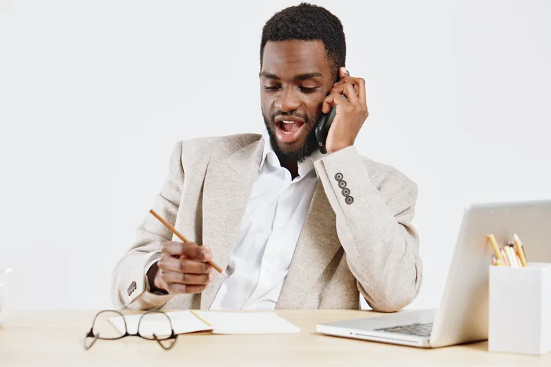 Professional man talking on phone, sitting at desk with laptop and glasses, in office environment. Business, communication, work, entrepreneur, workplace, technology, professional, seated, smiling.