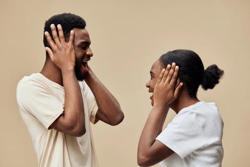Happy young couple joyfully playing with their hands over their ears, expressing fun and excitement against a soft beige background, ideal for emotional and lifestyle concepts