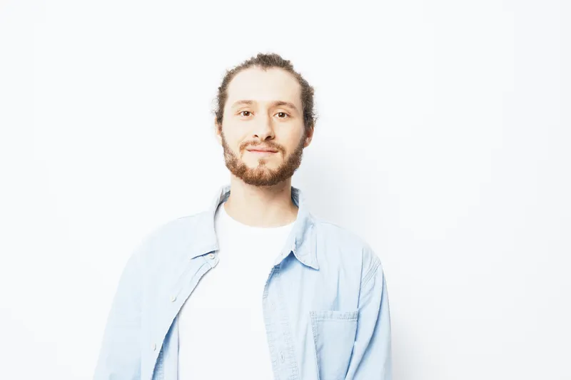 Smiling young man with a beard in casual denim shirt stands against a white background, radiating confidence and positivity, ideal for lifestyle and wellness themes