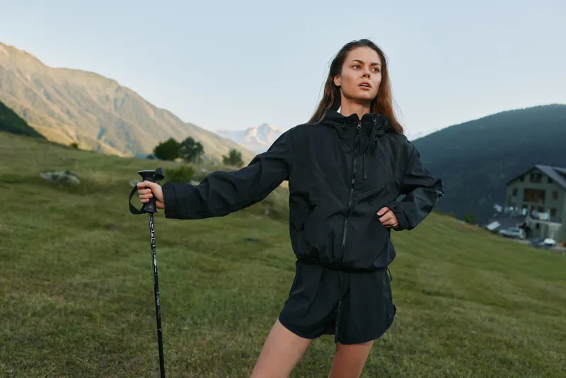 A stylish young woman stands on a grassy hillside with a trekking pole, wearing a black windbreaker and shorts, posing confidently against a scenic mountain landscape.