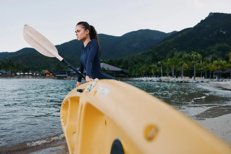 A paddler sits on a bright yellow kayak near a calm shore, gazing toward distant mountains as soft light highlights water, sand, and quiet shoreline.