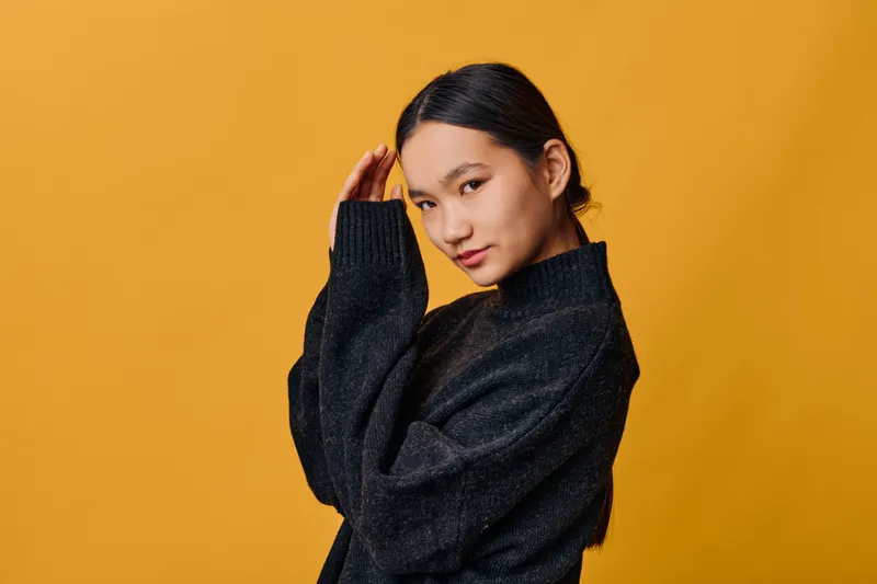 Young woman posing in black sweater on yellow background looking at camera with slight smile, confident posture and casual style indoors, studio portrait with soft lighting.