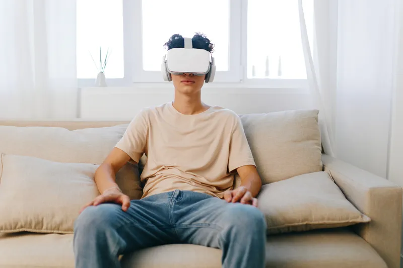 Молодой человек погружен в виртуальную реальность: он в VR-гарнитуре и наушниках сидит на уютном диване