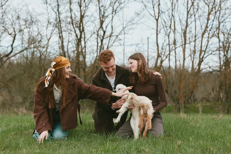 Three friends with a goat kid outdoors in a grassy field, smiling and interacting. Warm daylight highlights friendship, animals, rural charm, and rustic lifestyle, capturing a candid moment of