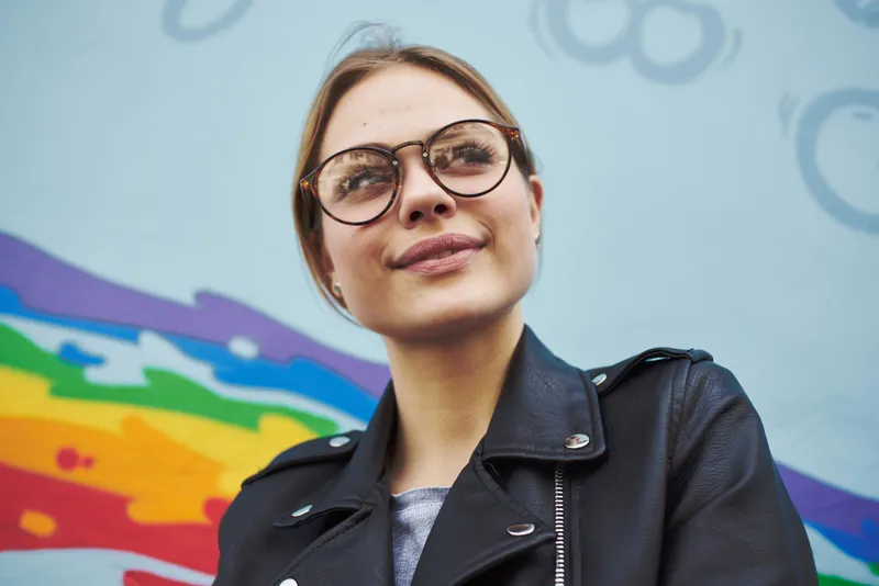 Счастливая женщина в кожаной куртке и очках стоит рядом с разноцветным граффити на стене
