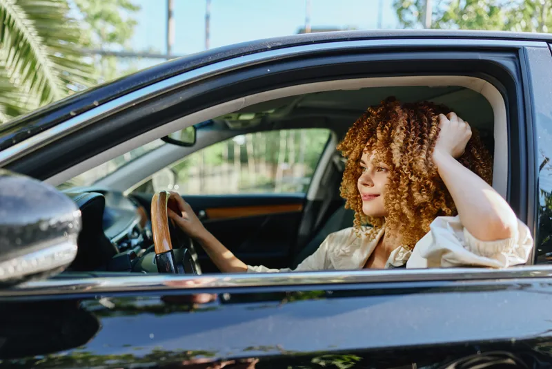 curly woman driving black car with relaxed expression in natural daylight, wearing casual light blouse, captured through open window in green outdoor setting