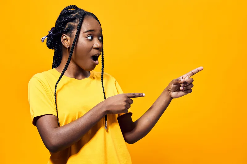 Surprised young Black woman in a yellow t shirt pointing to the side against a vibrant yellow background, expressing excitement and joy