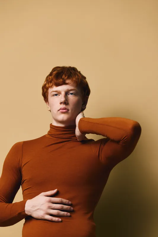 Young man with red hair wearing brown turtleneck posing with hand on neck and stomach against beige background. Portrait with serious expression and casual style.