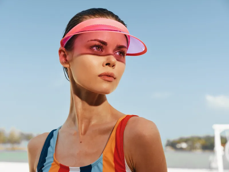Portrait of a young woman outdoors wearing a bright striped top and a pink visor, captured under a clear sky with soft sunlight and a confident, sunny vibe.