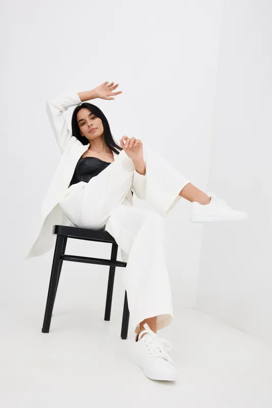 stylish woman dressed in white suit and black top posing on black chair in minimalistic white interior with casual white sneakers, contemporary fashion and relaxed mood