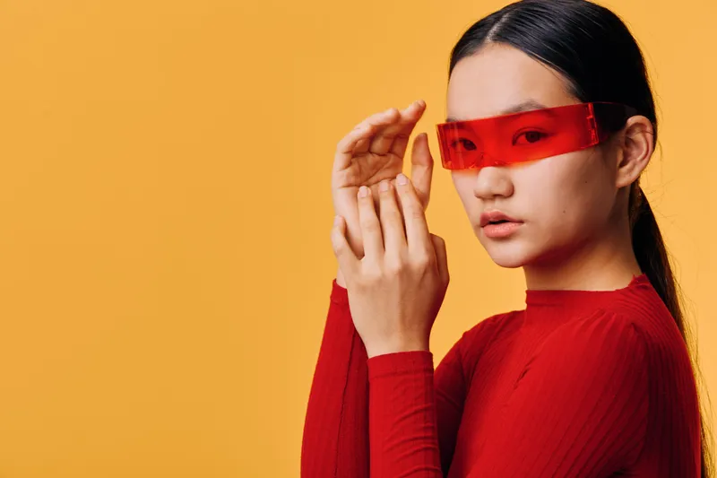 Young woman wearing red futuristic glasses and red sweater posing on orange background. Stylish female model with long black hair looks confident and modern in minimal fashion portrait.