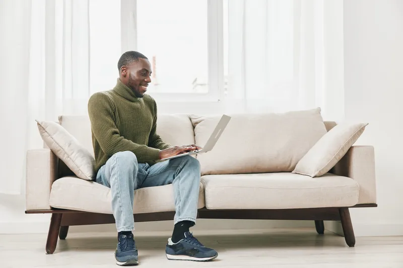 Young African man in casual wear happily working on a laptop while sitting on a cream colored sofa in a bright, modern room with natural light